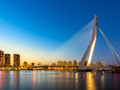 The Erasmus Bridge over a river at night, with city buildings and a skyline reflected in the water. Modern architecture and skyscrapers form the backdrop.