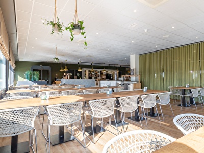 A restaurant with tables and chairs, a coffee table, and a vase, where large windows let in natural light.