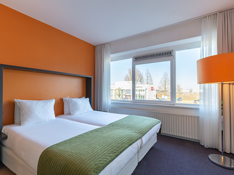 Comfort Twin kamer in Amrâth Airport Hotel Rotterdam met een groen sprei op het bed, een oranje muur en een oranje lamp.